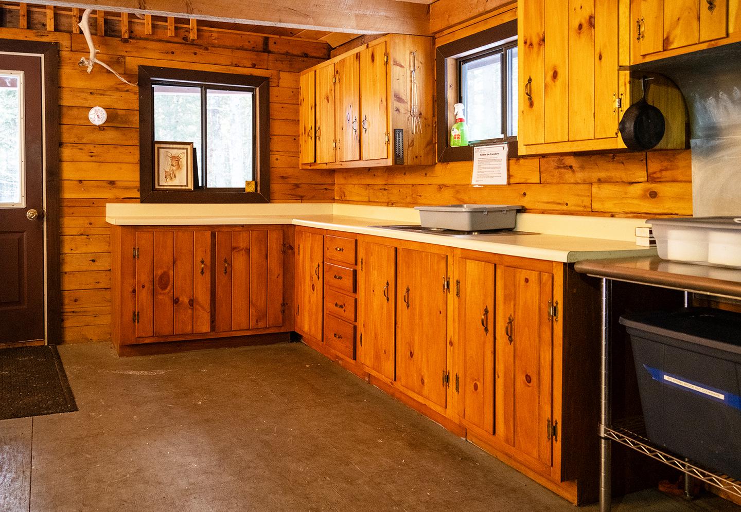 A kitchen space in a wooden cabin. A brown wooden door that leads outside is at the front. Big Spring Brook Hut has a large kitchen space and a door that leads to a porch outside.