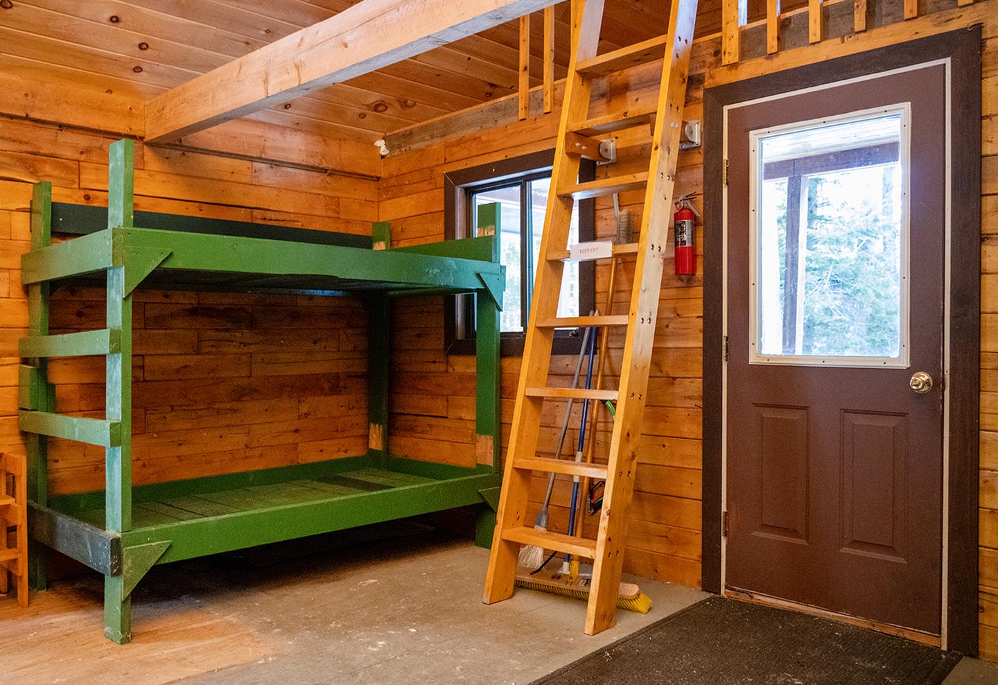 A wooden green bunk next to a wooden ladder (not for visitor use). A door with a window lets in light next to it.There are 4 bare wooden bunks inside. Remember to bring your own sleeping and camping gear!