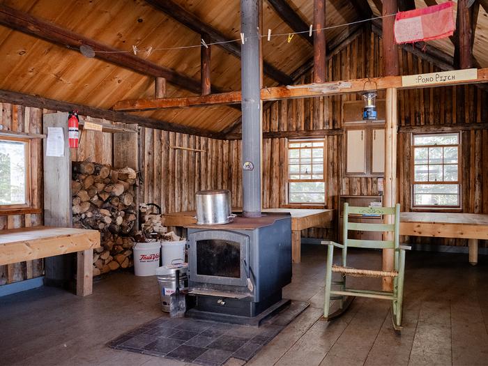 Inside a small wooden cabin. An old black woodstove is in the center of the common space. Platform bunks, neatly piled wood, and a rocking chair are in the same room. Three windows let in light.The spacious interior of Haskell Hut includes a woodstove and raised platform wood surfaces for sleeping. 