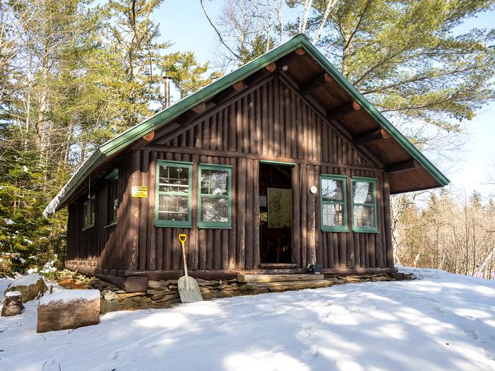 Haskell Hut on a clear blue sky day. Snow is on the surface of the ground and the door is open. Situated along the East Branch Penobscot River, Haskell Hut offers overnight accommodations for groups up to eight people in the winter.