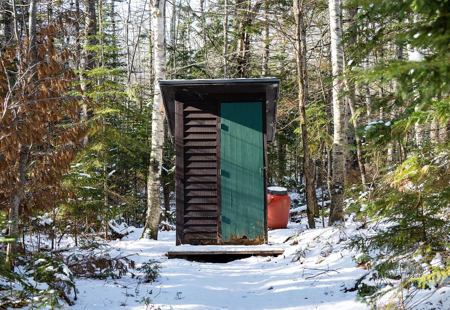 A tall wooden rectangular shack like structure with a green door. Snow is on the trail that leads to the single outhouse in the woods.An outhouse is available just outside of Haskell Hut.
