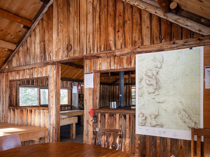 Inside of a wood cabin.  A large topographic area map hangs on the right side, and a dining table is hinted in the image. A wood wall made of separate logs separates this space and the living space behind it.Enjoy the shelter of Haskel Hut during the winter.