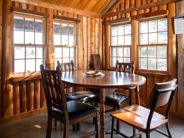 A wooden dining table sits in a corner with 4 small windows inside a wooden cabin. Games are placed on the table for visitor use.Enjoy the beautiful landscape from inside Haskell Hut during the winter.