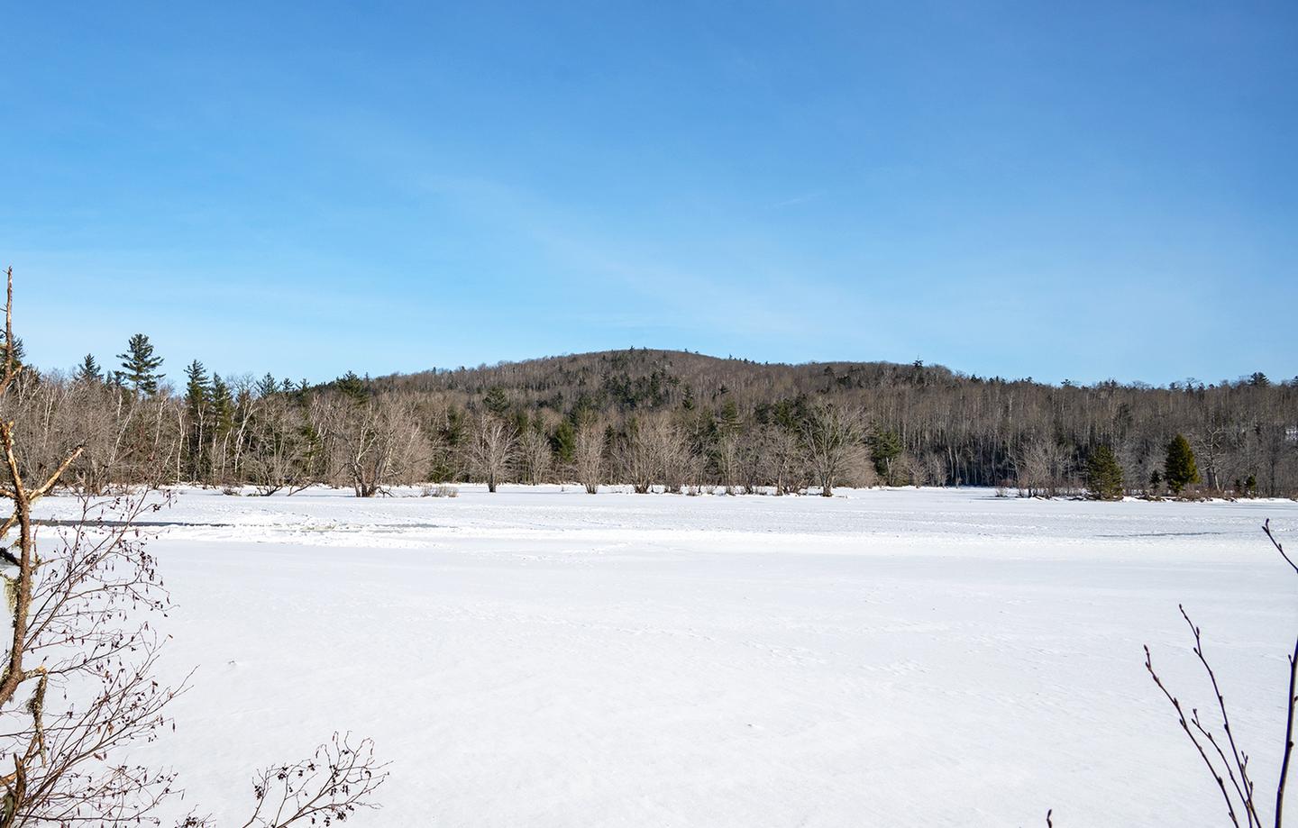 A winter landscape. Clear blue skies with a hill in the distance. Trees are bare. A frozen river with snow on top sits in the foreground.Enjoy views of the East Branch of the Penobscot River from Haskell Hut.