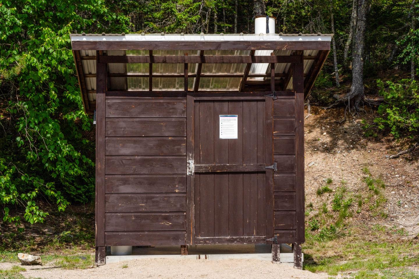 A brown wooden shack-like structure that shelters a pit toilet. The shelter has a leveled cement floor inside of it.A pit toilet is shared by all three campsites.
