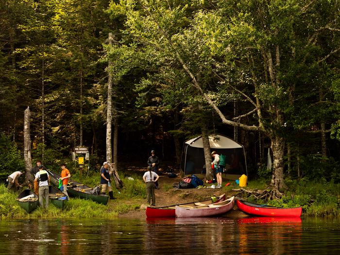 View of a campsite from the river. A group of campers and their canoes at the grassy and dirt bank of the river with their camping gear. The forest is shaded behind them with full green canopies.Big Seboeis Campsite can be accessed by boat.