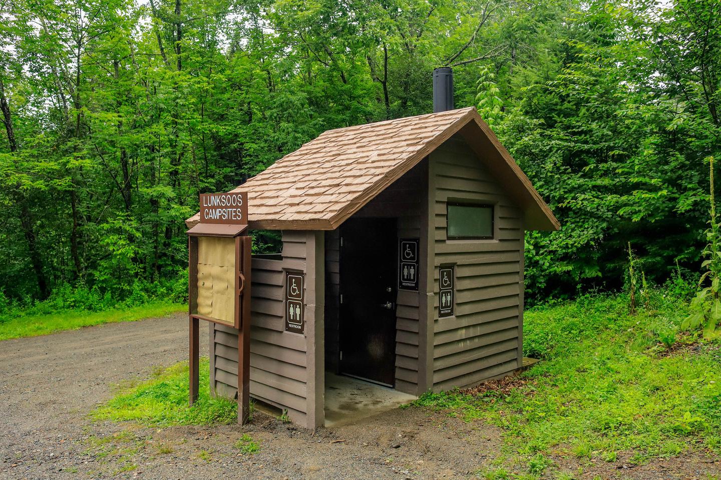 A small brown vault toilet building with an empty display sign against the left side of the building. The building is next to a gravel road with bright green grass on each side and tall trees.There is a vault toilet for use near the entrance of the campground parking lot.