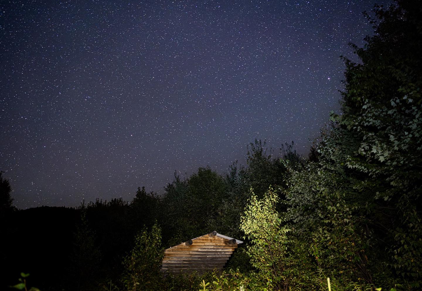 An expansive dark night sky with many stars above a small wooden structure. The silhouette of the forest surrounds the wooden structure.Lunksoos Lean-to is a wonderful location to stargaze and enjoy the night sky.