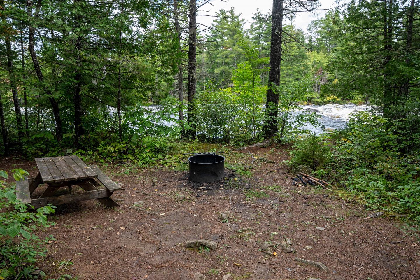 A campsite next to a river. The campsite has a wooden picnic table and a metal fire ring. The ground is made of dirt and gravel with some uneven surfaces. Brush and tall trees surround the perimeter of the campsite.Enjoy the views of the East Branch of the Penobscot River from this campsite.