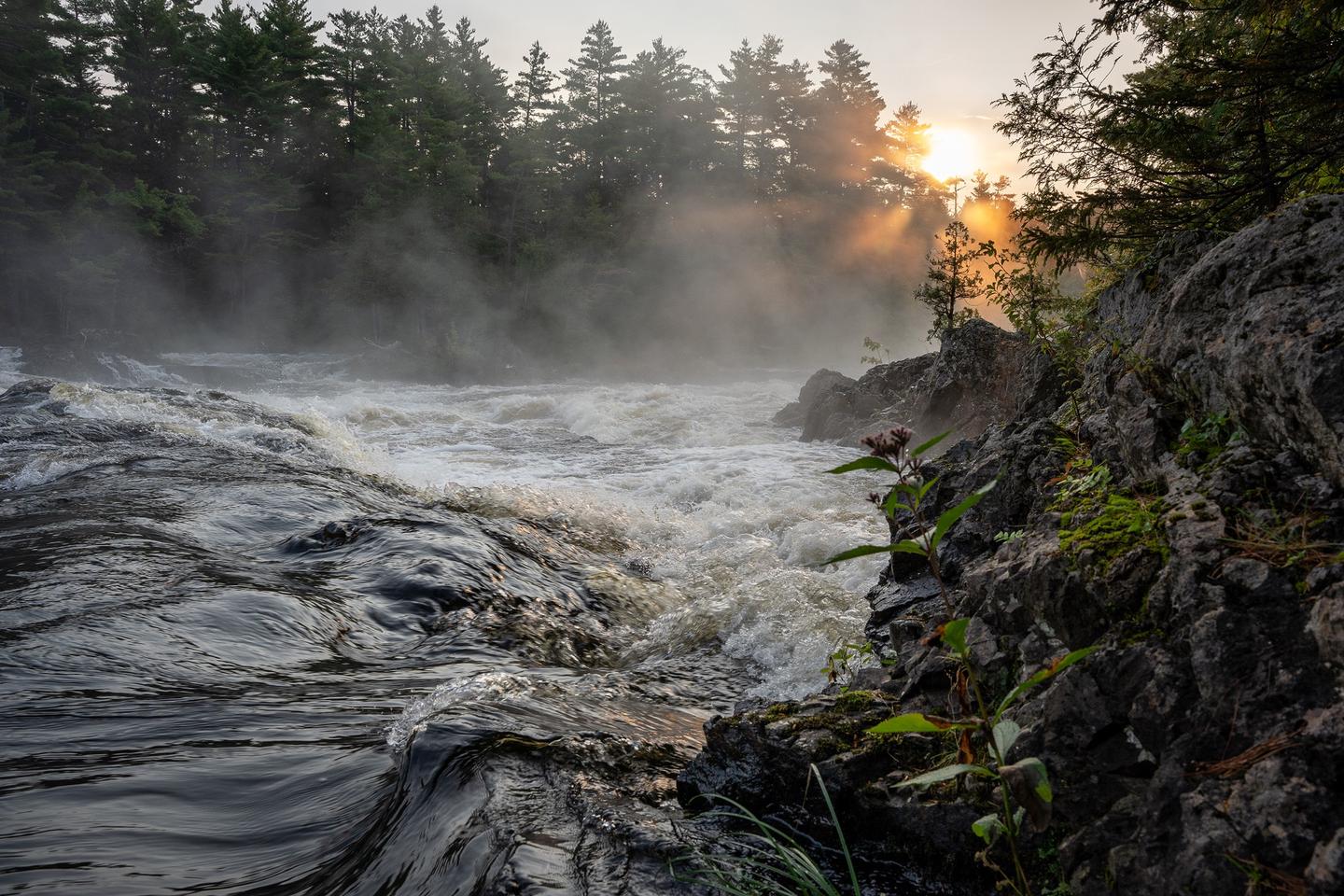 A close up view of a rushing river rapid with the forest in the distance and rocks to the right.Enjoy the beautiful sounds and views of the East Branch of the Penobscot River from this campsite.