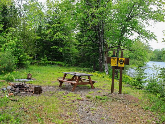 A gravel and dirt campsite above a bank of a river surrounded by tall trees and grass. The campsite has a metal fire ring on the left, a brown wooden picnic table, and a brown wooden sign for Stair Falls Campsite and portage. The sign has two yellow signs reminding visitors about registering fires with the state of Maine.This campsite has a wide open grassy area with a view of the river.