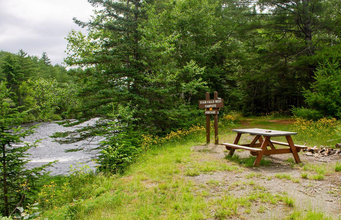 A river runs below a gravel campsite to the left. A brown wooden sign shows Stair Falls Campsite and Portage. A wooden picnic table is available on the right for visitor use with firewood next to it. A dirt trail extends into the shaded forest behind the campsite..Stair Falls Campsite is located on the East Branch Penobscot River near the portage trail.