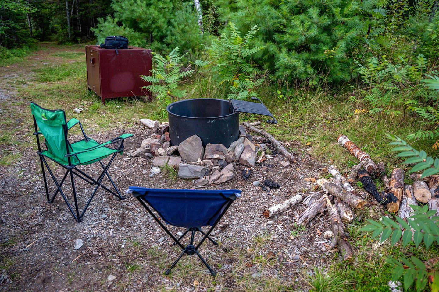 A metal fire ring at a campsite during the daytime. There are two camping chairs next to the empty fire ring with large rocks and firewood on the ground around it. A brown metal food storage box is adjacent to the fire ring. Green grass, brush, and trees border the campsite.Remember to call Maine Forest Service at 207-435-7963 for a fire permit.