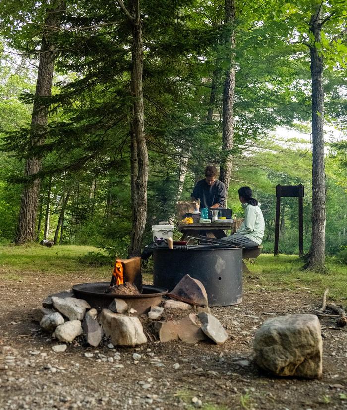Campers with a morning fire. Wood burns in a metal fire ring as two campers prepare a meal on a wooden picnic table behind it.Upper East Branch Campsite offers a wide space that can fit multiple tents.