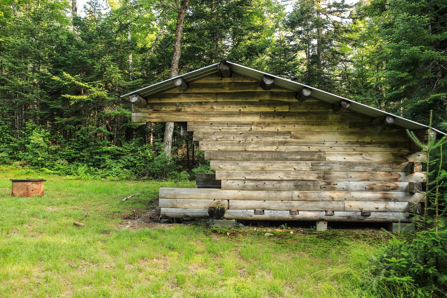 Facing the side of the lean-to. A raised wooden structure made of logs with an angled roof covering. A fire ring is available for use! Remember to contact the Maine Forest Service at 207-435-7963 before your trip. 