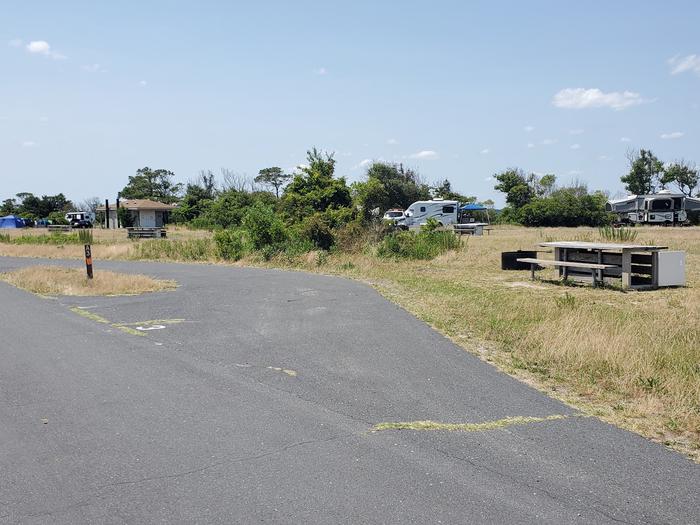 Front view from main road of bayside site A3 in February.  Image shows the pull-through drive-in site with a wooden picnic table and black metal fire ring to the right of the campsite.  Bathrooms within view to the left of the campsite along with other campers.Bayside A3 - June