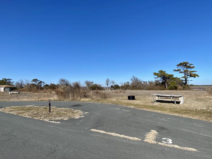 Front view from the main road of bayside site A3 in February with a view of the paved pull-through drive-in site. There is a wooden picnic table and black metal fire ring to the right of the site.  There is a white spray-painted 3 to the right at the entrance of the pull-through site.Bayside A3 - February