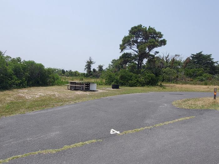 Bayside site A4 in July with a view from the left on the main road of picnic table and fire ring with trees and brush to the right.  Partial view of pull-through drive-in site.Bayside A4 - July