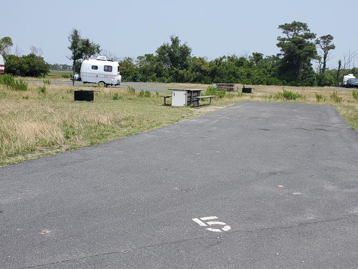 Bayside Site A5 in July. View from the main road and shows the entire back-in drive-in site.  Wooden picnic table and black metal fire ring to the right of the campsite.  Another camper's trailer within view behind the campsite. There is a white spray-painted 5 on the pavement at the entrance to the campsite.Bayside A5 - July