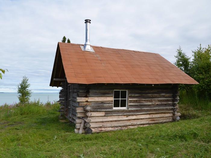 A wooden cabin with a window and a stove pipe coming out of the roof in front of a lakeBig Bay cabin.