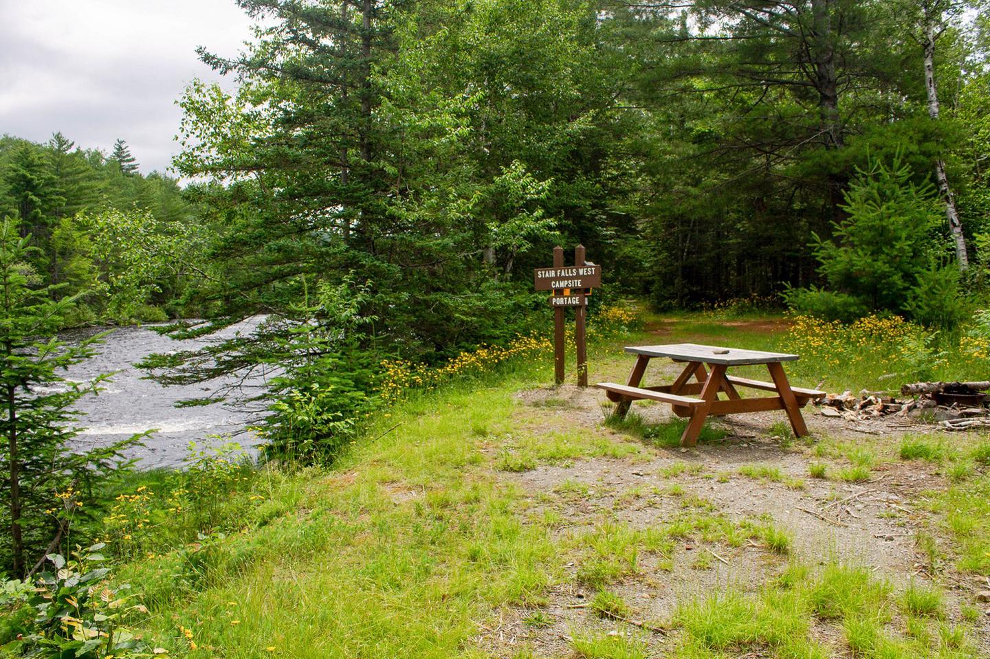 A gravel and grassy campsite to the right of a river. The campsite has a wooden picnic table, a brown wooden sign, and a trail that leads into the woods with tall dense green trees.Stair Falls campsite along the East Branch Penobscot River has a beautiful view of the river.