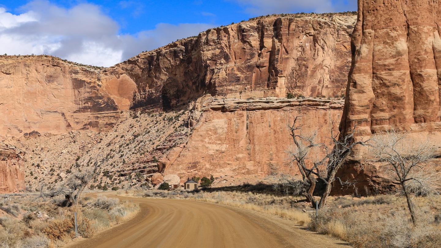 A dirt road cuts through a wide canyon of orange cliffs with a bathroom in the distance.The alcove area, sites 18-21.