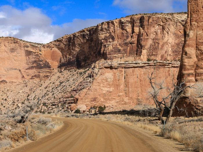 A dirt road cuts through a wide canyon of orange cliffs with a bathroom in the distance.Alcove area at Buckhorn Draw campground