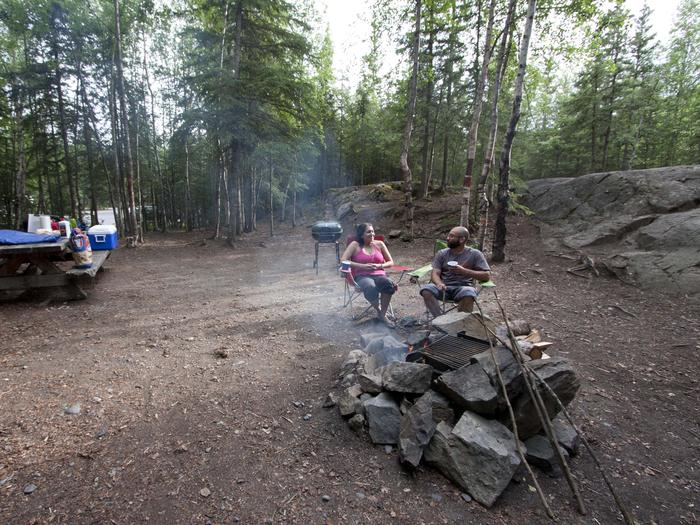 Campers relax in campground next to smoking campfire, background of forest and a picnic table holding supplies.Rustic campsites are surrounded by spruce and alders, providing relaxing dry camping opportunities for campers.