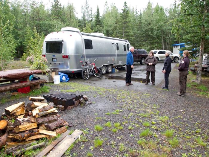 People stand in front of a silver camper trailer with firewood stacked in the foreground next to a campfire ring.Campground hosts are available from May through September to assist campers with firewood sales, same day reservations and work with refuge rangers to provide a safe, enjoyable camping experience.