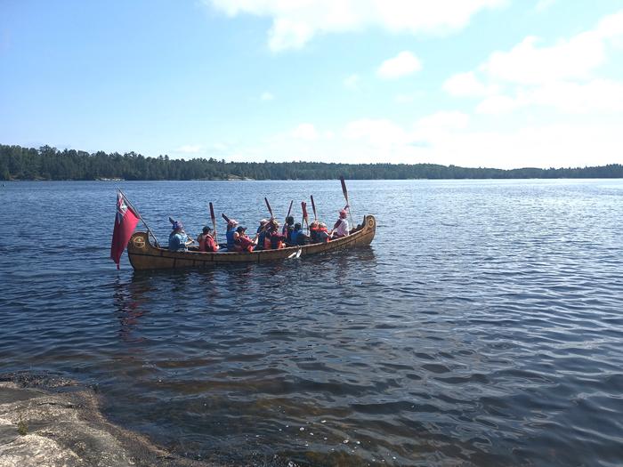 Park visitors paddle a 26-foot-long north canoe on Kabetogama Lake near the Ash River Visitor Center with a forested shoreline in the background.Park visitors enjoy learning about the life of a Voyageur aboard a 26-foot North Canoe.