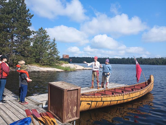 Rangers dressed as French voyageurs stand in front of visitors on a dock next to a 26-foot North Canoe.