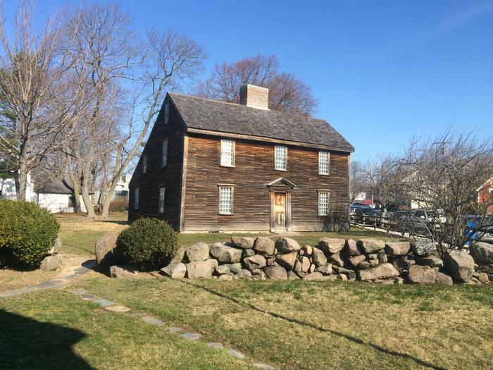 A New England "saltbox" style home with brown wooden siding. In the foreground, there is a low stone wall, two large shrubs, a stone pathway, and grass. There are trees with no foliage behind the home.The John Adams Birthplace