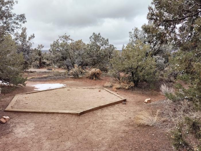 A small tent pad surrounded by trees and shrubs.Site 2 has a tent pad, picnic table, and metal fire ring. The site is surrounded by pinon-juniper forest.