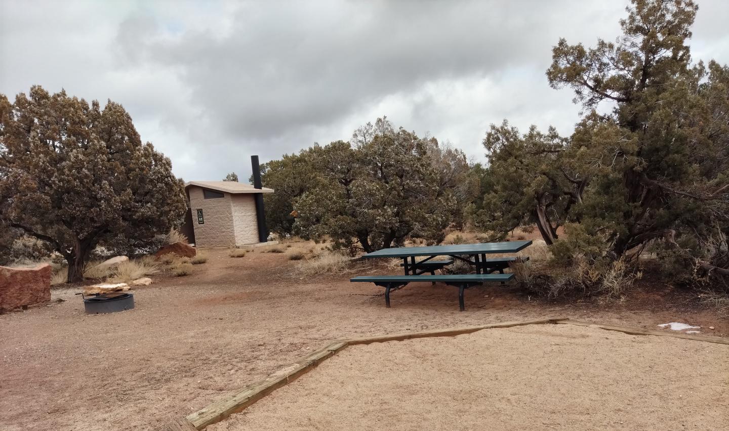 Picnic table at Site 11. Site 11 has a tent pad, a picnic table and a metal fire ring. The site is surrounded by a pinon-juniper forest.