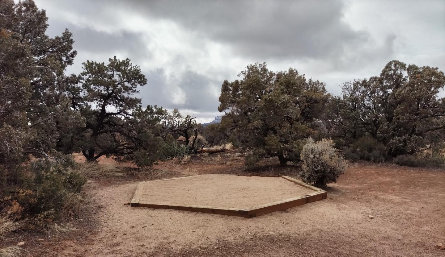 Small tent pad at Site 12, with scrubs and trees in the background.Site 12 has a tent pad, a picnic table and a metal fire ring. The site is surrounded by a pinon-juniper forest.
