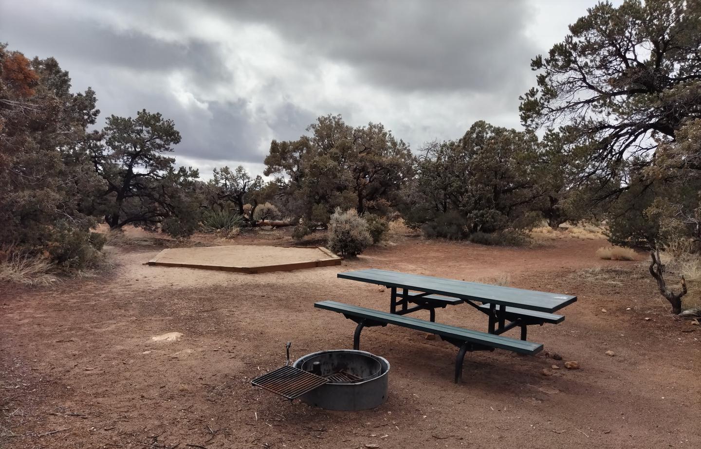 Picnic table, metal fire ring, and tent pad at Site 12.Site 12 has a tent pad, a picnic table and a metal fire ring. The site is surrounded by a pinon-juniper forest.