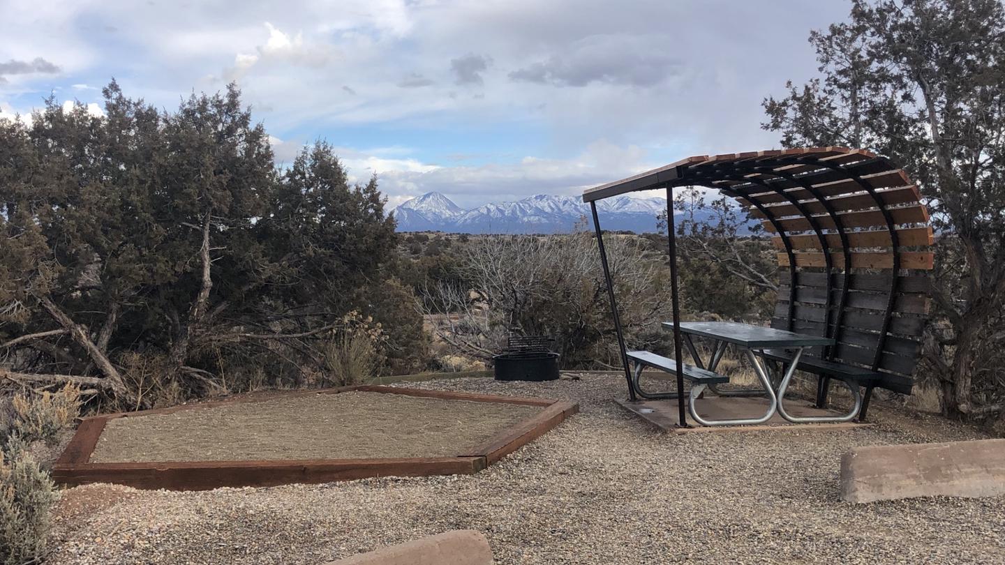 A tent pad and covered picnic table in front of a mountain range.The Hovenweep campground has views of mountains and canyons and is surrounded by desert vegetation. 