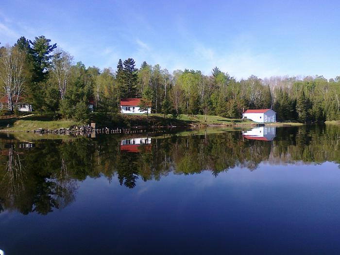 White cabins with red roofs nestled along a shoreline of trees reflect perfectly off of a blue, calm lake surfaceReflection of Hoist Bay cabins on a calm day.