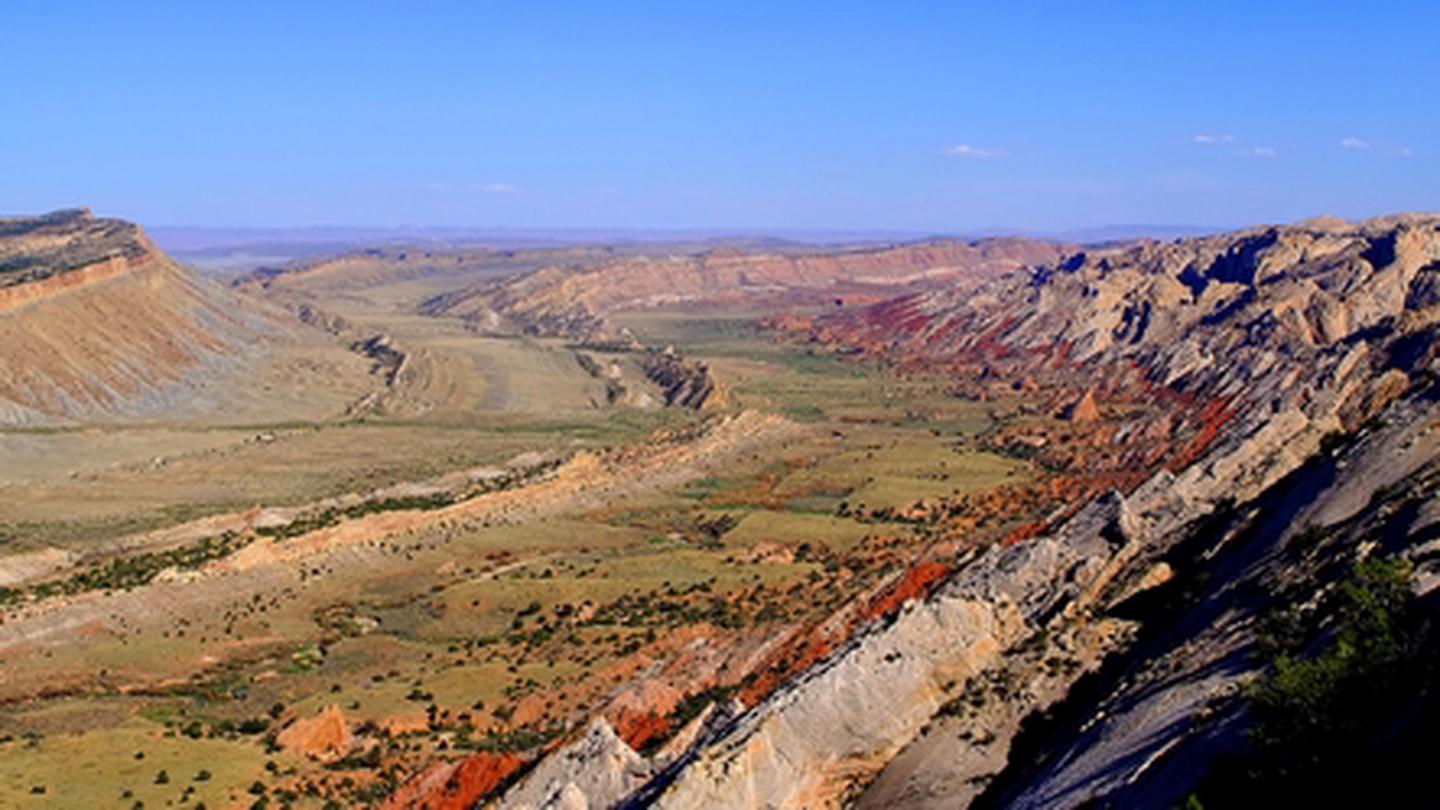 A green valley, a brown ridge cuts through it. Red and brown cliffs slope up on the right side of image. The sky is a bright, cloudless blue.The Strike Valley shows colorful layers in the Waterpocket fold.