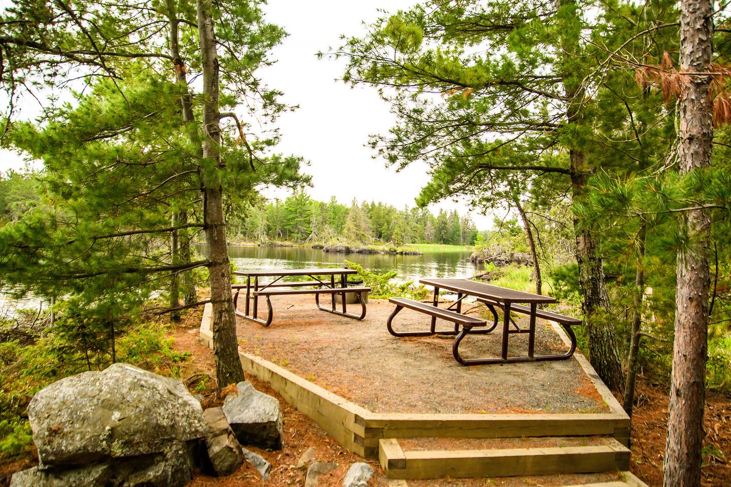 Picnic tables and fire ring between pine trees with lake in backgroundDay use area at Oveson's Fish Camp