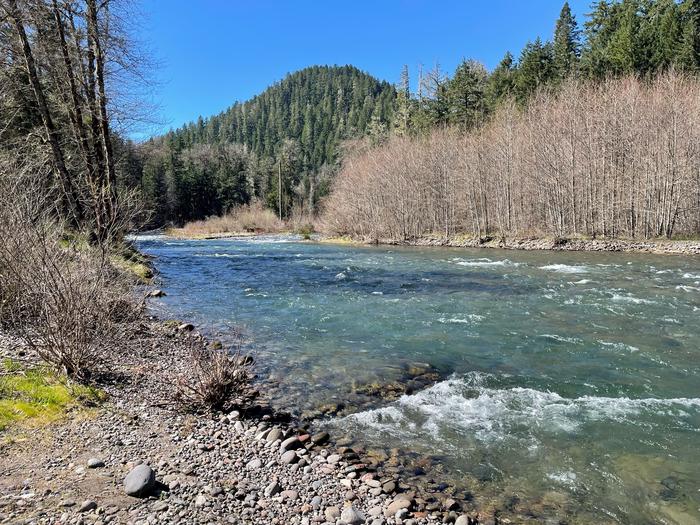 A rocky beach leading into a blue river. Small tree covered peaks can be seen on the other bank.The Middle Fork River runs close to the Sand Prairie Campground, with the Middle Fork National Recreation Trail starting in the campground and continuing along the river for 30 miles.