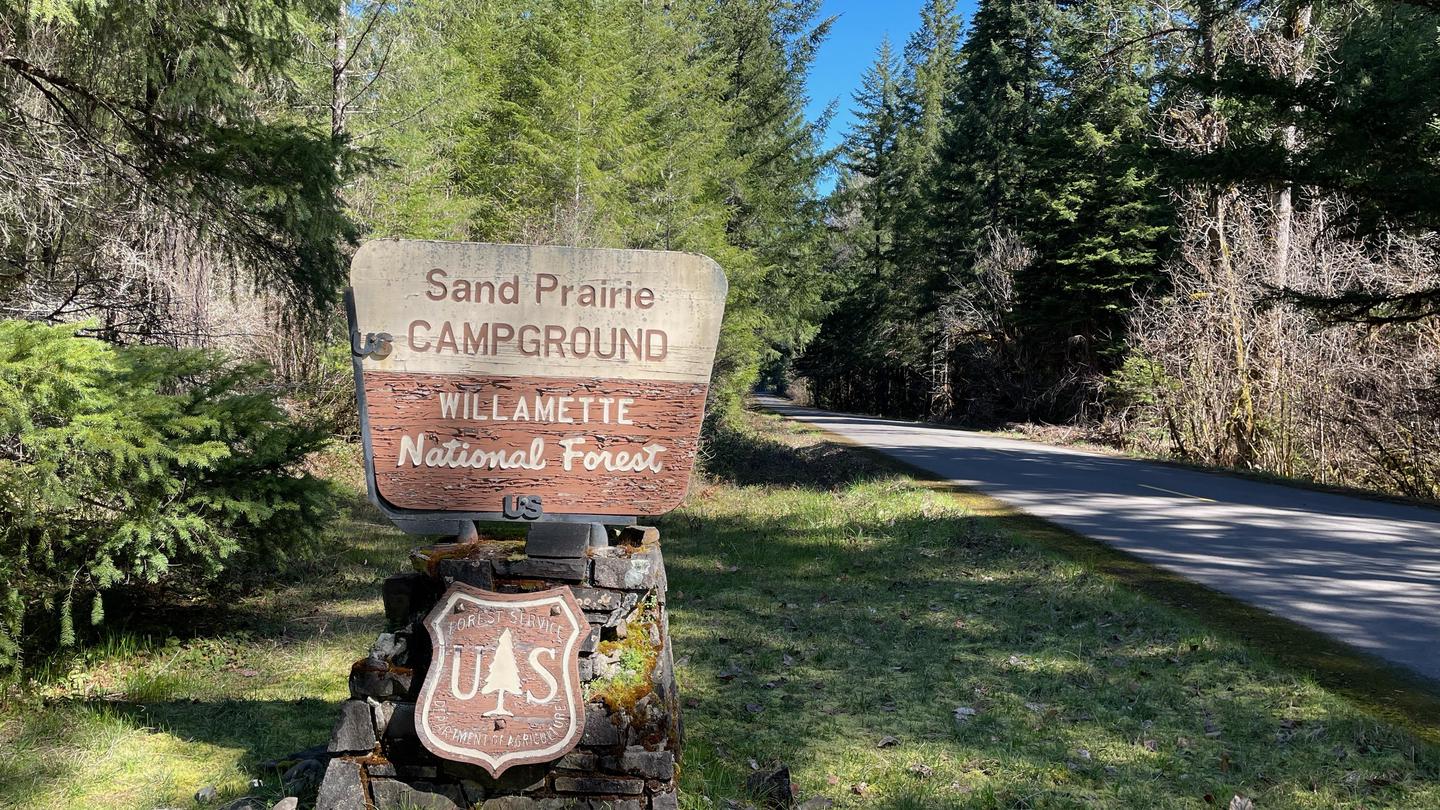 A wooden sign on a stone base. The sign is brown and cream colored and reads "Sand Prairie Campground"The entrance sign for Sand Prairie Campground can be seen easily from Forest Service Road 21 and guides you into the campground.