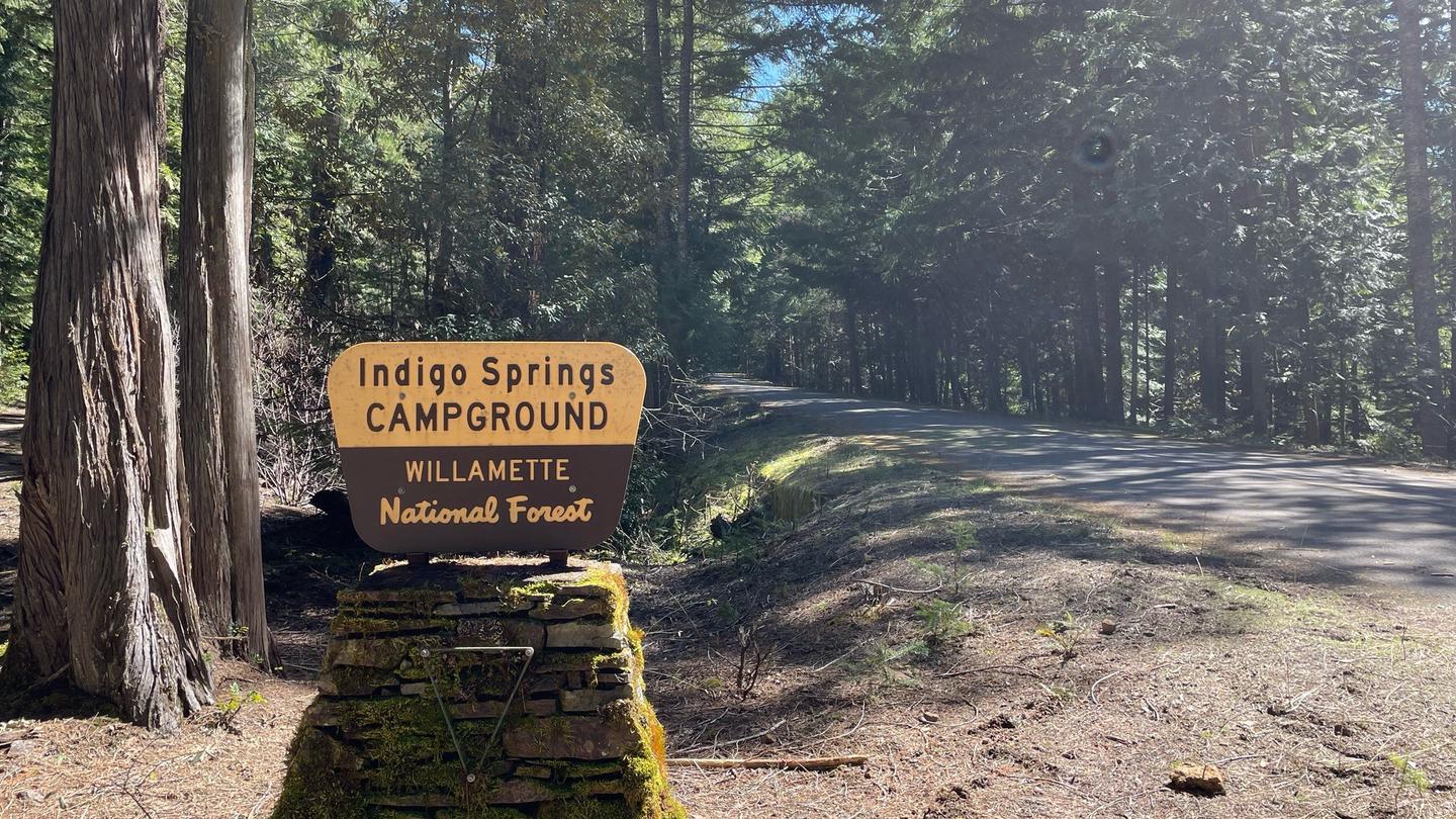 A cream and brown sign on a stone pedestal. The sign reads "Indigo Springs Campground"The entrance sign to Indigo Springs can be seen easily from Forest Service Road 21 and leads directly into the small campground.