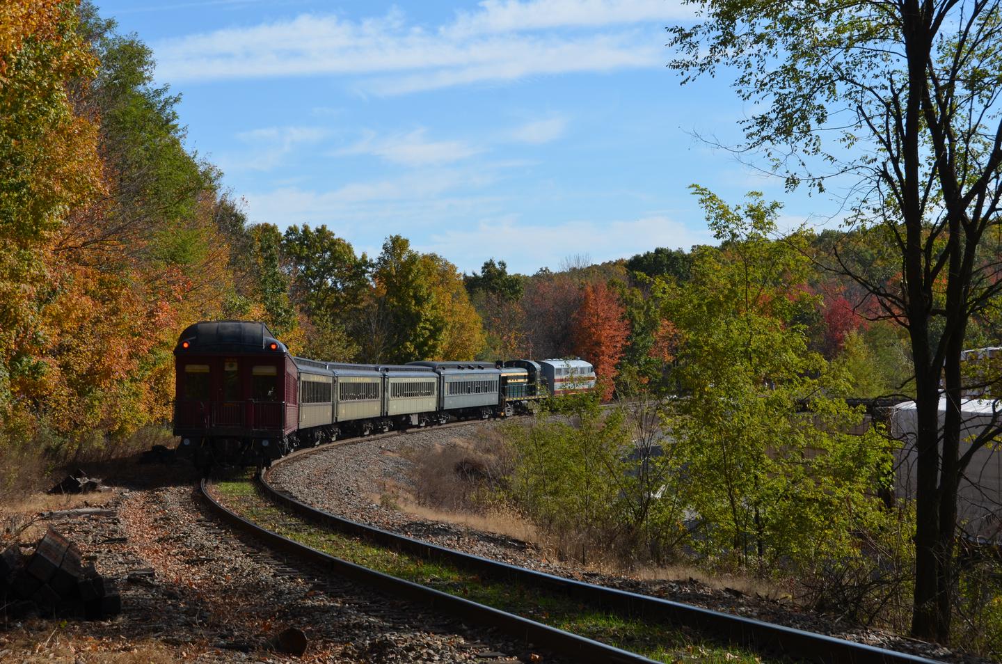 The image is that of a train pulling away.  There are two diesel electric engines and five passenger coaches.  train is surrounded by trees.  The leaves are in fall colors of red, orange, yellow, and green.  Train leaving Cresco Station.