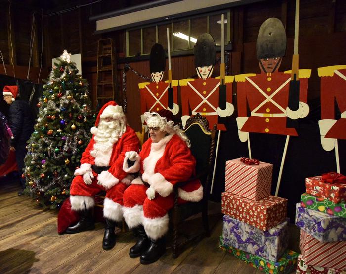 Santa Claus and Mrs. Claus are seated next to a decorated tree.  The tree is to the right of Santa and Mrs. Claus.  There are wrapped presents to the right of them.  Behind them stand life size wooden toy soldiers.Santa and Mrs. Claus