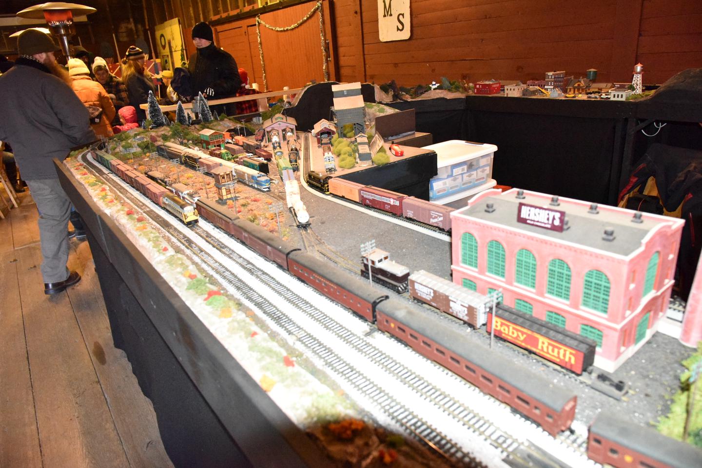 There is a model train display.  There are many buildings, tracks and model trains on the  display.Model Train Display