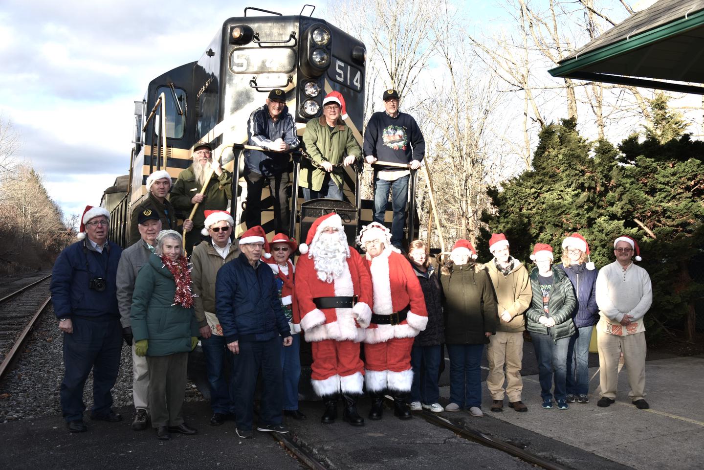 Santa and Mrs. Claus are standing in front of a diesel electric locomotive.  This is a staged photo.  They are surrounded by fifteen volunteers and two park rangers.The Moscow Holiday Express Team