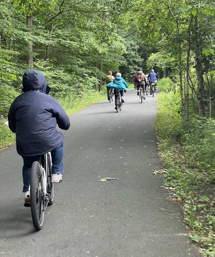 There are several people riding bicycles and a paved train through a wooded area.Biking the Lackawanna River Heritage Trail