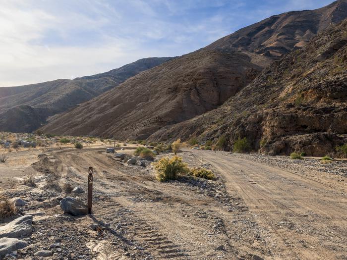 Gravel dirt road with pull-through campsite loop, brown post on ground labelled C-5, hillside backdrop with blue skies.Site C-5 in Cottonwood Canyon Road.