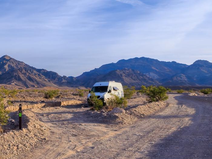 White camper van in a gravel dirt road with pull-through campsite loop, brown post on ground labelled E-1, hillside backdrop with blue skies.Site E-1 along Echo Canyon Road.